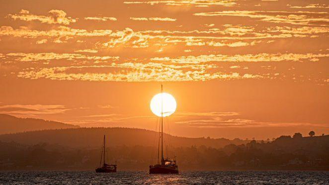 The sunsetting over the sea. Two boats are sailing. The sky is orange.