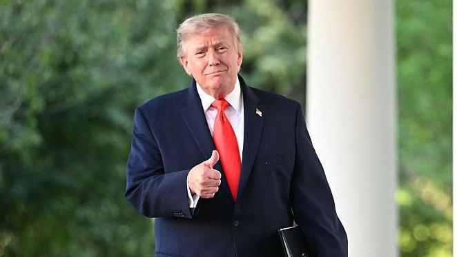 Donald Trump walking and giving a thumbs up. He is wearing a navy suit, white shirt and red tie.