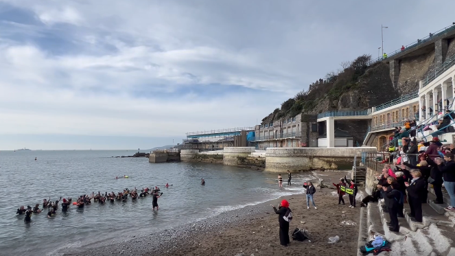 About 50 members of the Rock Choir in the sea. The group are facing the shoreline. About 30 people are standing on steps on the beach as they support the choir group for the sing and swim event. 