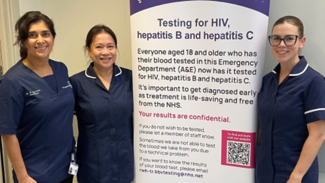 Three women in blue hospital uniforms standing beside a Hepatitis information sign. It outlines details for the testing scheme headlined "Testing for HIV, Hepatitis B and Hepatitis C". All three women have long dark hair, pulled back in pony tails and one has glasses. All three smile at the camera.