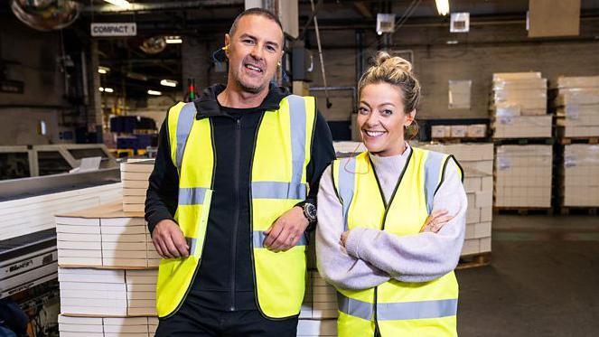 Presenters Paddy McGuinness and Cherry Healey are wearing yellow fluorescent vests and are standing in a factory setting surrounded by crates of paper