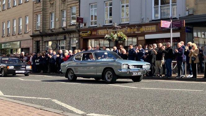 A light and dark coloured Capri drive past applauding crowds on the roadside.