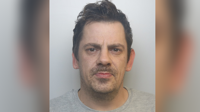 A custody mugshot of Mark Roberts. He is wearing a grey jumper and has short dark hair and dark eyes with stubble and is looking into the camera.