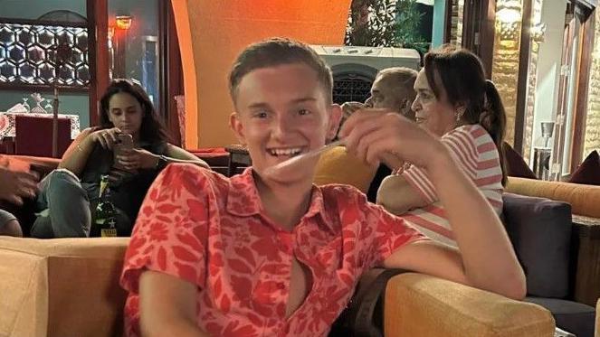 Jack, sitting in a public place, in a red shirt, smiling to the camera before his accident