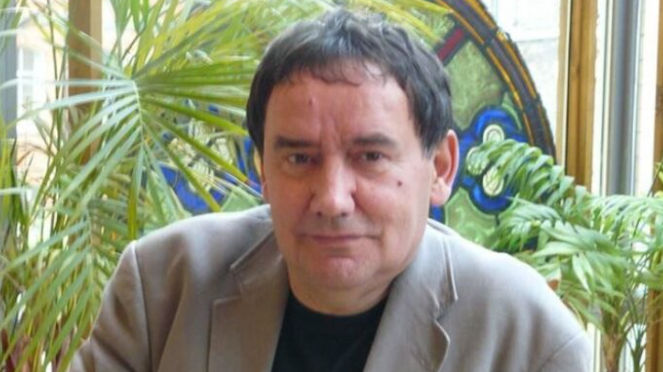 A handout of Graeme Phillips looking into the camera. He has dark black hair and is wearing a light jacket and black top. He is sitting in front of several plants.