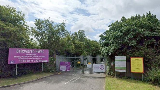 Grey gates closed across the entrance to a recyling centre. A purple sign indicates that it is "Brixworth HWRC". There are other signs either side of the entrance, with trees and hedges behind.