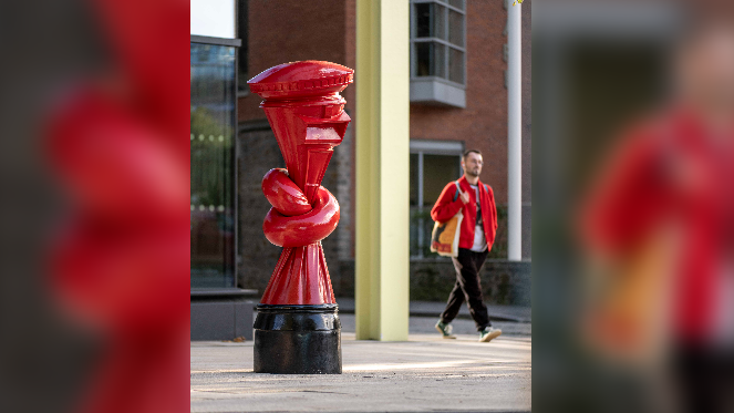 A man wearing a red jacket holding a canvas bag walking past a red post box that has been tied into a knot