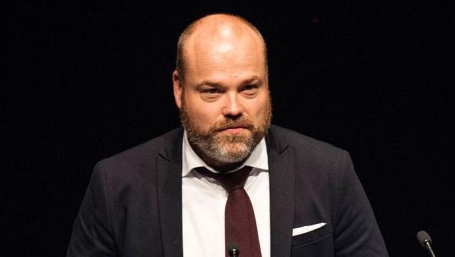 Anders Holch Povlsen is a bald man with a brown beard. He is wearing a dark jacket, white short and brown tie