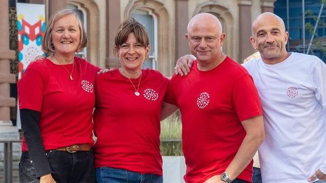 Two women stand in a line next to two men. The two women and one of the men all wear the same red T-shirts. The other man on the end wears a white long sleeved T-shirt. One of the women has bobbed blonde hair while the other has brown hair that has been tied up. Both men are bald. 
