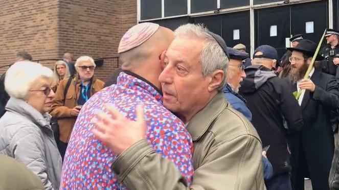 Two men, in traditional Jewish Kippah caps, embrace outside a synagogue building within a small crowd.