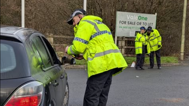 A male police officer in a high-visibility jacket leans in as he talks to someone in a dark car parked up as part of a drink drive campaign. In the background two other officers are talking in front of a large sign and some trees.