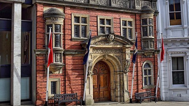 The legislative chambers, a red brick building with a grand brown entrance, there are four flag poles in front.