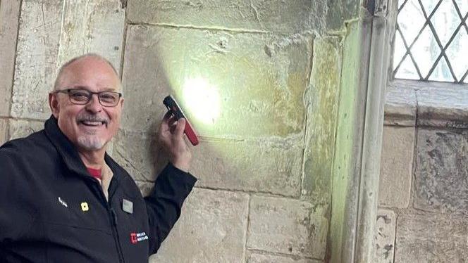 Rick Berry is standing next to a wall holding a lit torch against it. He is smiling and is wearing glasses.