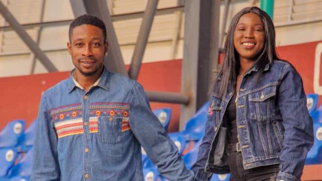 A male and female dressed in denim jean holding hands and looking into the camera while standing in part of a stadium with blue plastic seats in the background