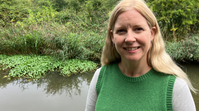 Karen Lewing has long, blonde hair and is wearing a green tank top. She is standing by a canal in Worcester with the invasive plant in the water behind her, which is growing like a green carpet against the banks of the canal