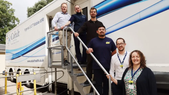 A group of professionals - men and women - stand on the metal staircase of a portakabin containing an MRI scanner. They are all smiling.