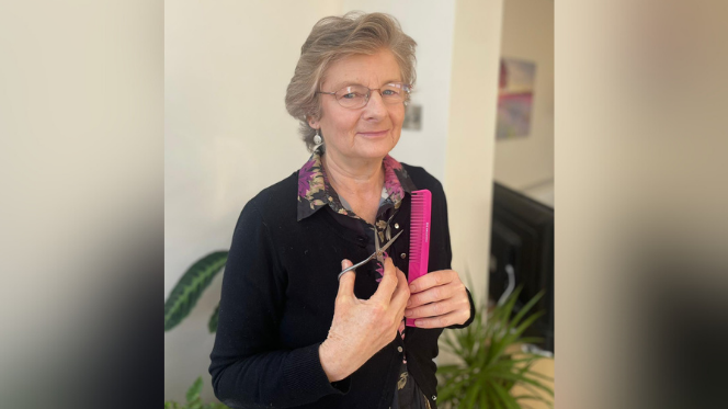 Petition initiator Jenny Wells. She is smiling for the camera and holding a magenta comb and a pair of scissors. There are plants in pots behind her.