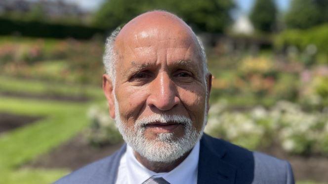 Mohammed Arshed has light brown skin with balding grey hair and a grey beard. He is wearing a grey suit and flower beds can be seen behind him