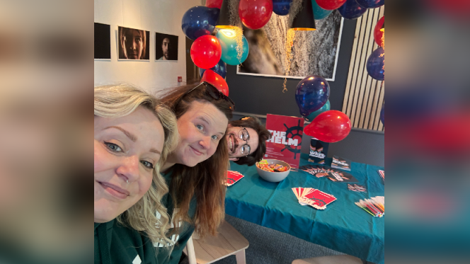 Two women and a man peer into the frame from the side, with a table at the centre of the image surrounded by red and blue balloons and leaflets advertising The Helm.