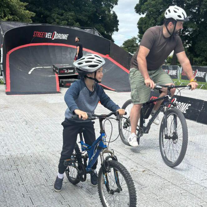 A man and a boy, both wearing casual clothes and helmets, ride bikes on a temporary cycle track. Behind them is a banked curve - a smaller version of those seen in velodromes - which is coloured black with red trim. It features the slogan "Street Velodrome". 