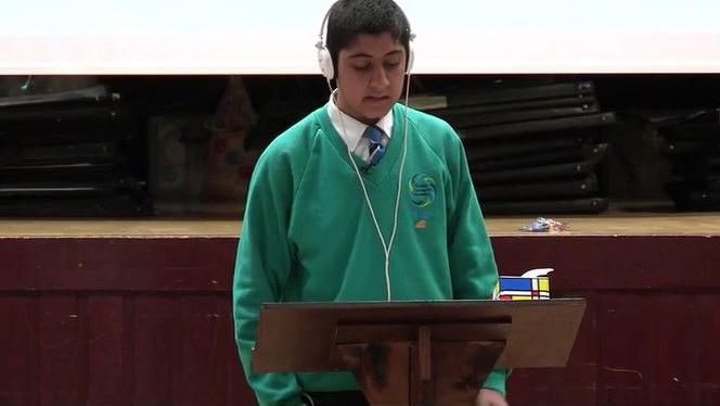 A Asian teenage boy wearing a green school jumper wearing earphones while standing at a lectern