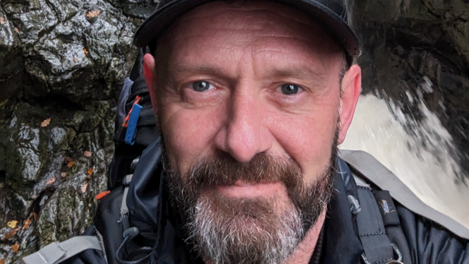 A man with a beard and black cap wearing a black coat and backpack, standing in front of rocks and a waterfall