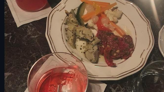 A glass of red liquid with ice sits on a marble table. Next to it is a plate of pickled vegetables, artichokes and sundried tomatoes.