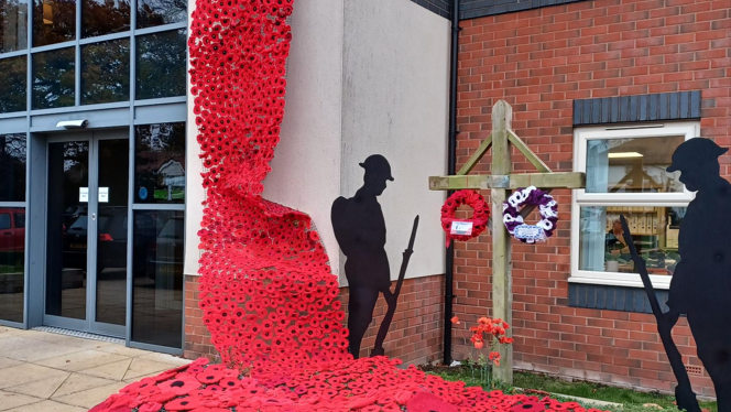 A stream of poppies fixed to the side of a building's entrance with two soldier silhouettes and a wooden cross beside it