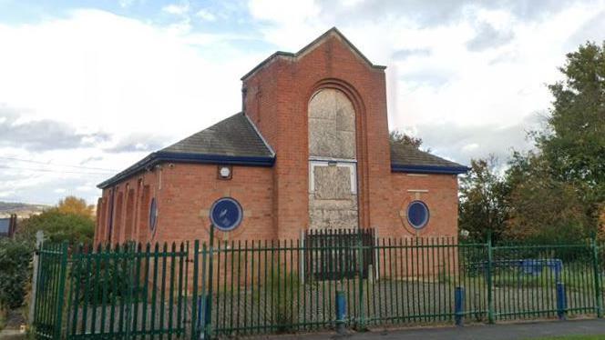 A red brick building, some windows covered by boards, and surrounded by a green metal fence