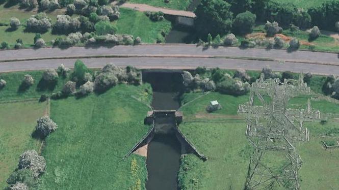 An aerial image of the gate, which sits between the riverbanks next to green fields. A road can be seen stretching from the left to the right of the photograph.