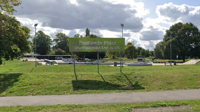 A green sign on a verge read "Radlands Plaza - Northampton Skatepark". The ramps etc., are behind the sign. There are lamp-posts and trees around the skatepark.