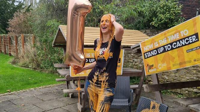 Faye Louise after being gunged with 15 litres of orange gloop in the garden of the Red Lyon pub in Slinfold. She is stood laughing, covering in orange gloop. 