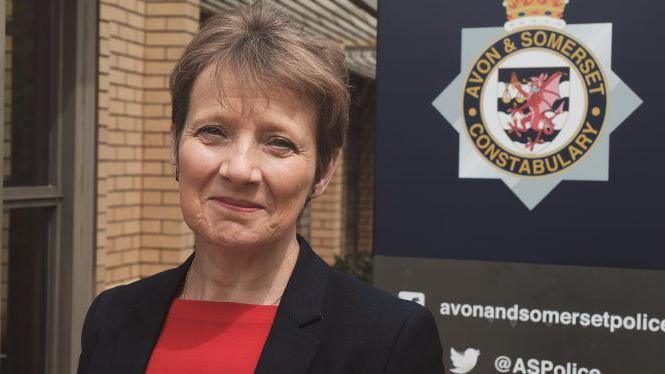 Clare Moody wearing a red top and black blazer. She has short brown hair and is smiling at the camera, standing in front of a sign that says Avon and Somerset Police Constabulary 