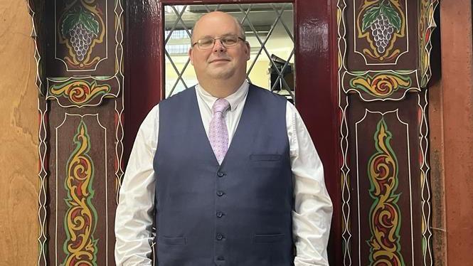 The director of the museum wears a waistcoat, tie and glasses and stands in front of a brightly decorated doorway