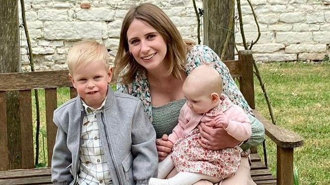 Kirstie is smiling at the camera with a baby on her knee and a little boy standing next to her. She is sitting on a wooden bench and is wearing a colourful top and has shoulder-length blonde hair. The baby is wearing a flowery dress and pink cardigan and the little boy is wearing a checked shirt and a grey jacket.