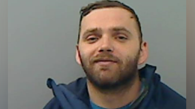 A man with short brown hair, a brown beard, smirks at the camera, in a police mugshot. The background is a painted grey wall and he is wearing a blue t-shirt and navy jacket.