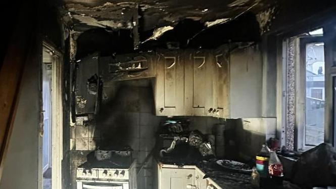 A burnout kitchen, with smoke and flame damage. The ceiling is black, with material falling down, a burnt white cooker, burnt cooking surfaces and damaged kitchen cabinets. A widow is opened and the glass is cracked. 