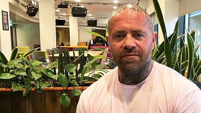 A man with a shaved head with tattoos and a dark beard. He is wearing a pink T-shirt and is sitting in the foyer of a BBC studio with plants behind him.