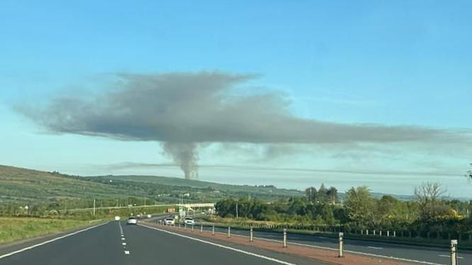 a large plume of smoke above fields. In the foreground is a dual carriageway