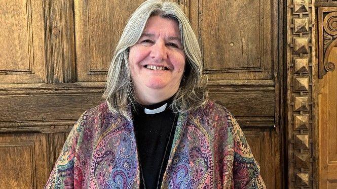 Bishop Worsley with long greying blond hair, a paisley patterned jacket with elements of violet, gold and green and a black clerical shirt with white collar.