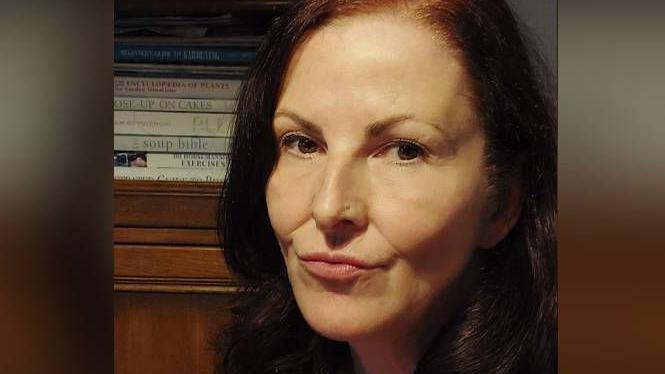 A woman with dark red hair, wearing a black cardigan over a read and black striped top looks into camera as she sits in a dimly lit room.