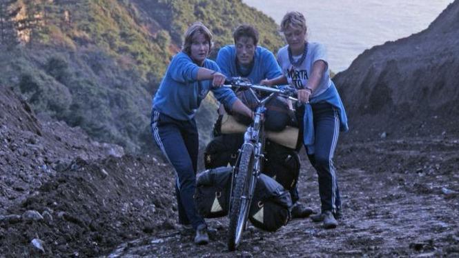 The three friends are pushing a bike loaded with bags up a steep, muddy slope. Craig is in the middle, and looks exhausted. Behind them are more steep slopes.