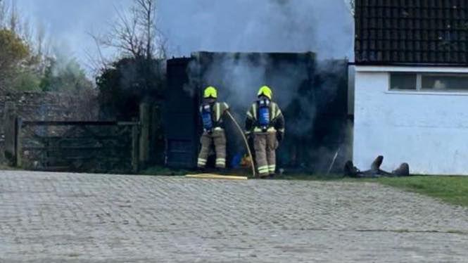 Two firefighters are stood outside of the container as they try to tackle the flames. Smoke is coming out of the container. The firefighters are wearing fire safety gear and yellow helmets. They are using a hosepipe to put flames out.