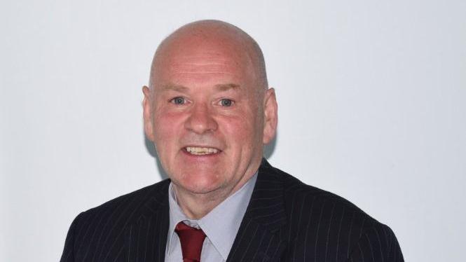 Labour Councillor Robert Moran in the middle of the picture taken from the shoulders up. He smiles at the camera whilst wearing a red tie with a grey shirt. His suit jacket is black with pinstripes. 