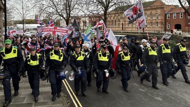A line of police in neon vests in front of a crowd of people holding different variations of the Union Jack, including some with slogans like "Unite the Kingdom" and . A row of red brick buildings can be seen in the background.