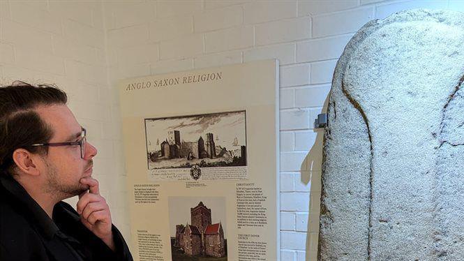 A man with glasses looks at a piece of art. Next to it is a board that says 'anglo saxon religion' 