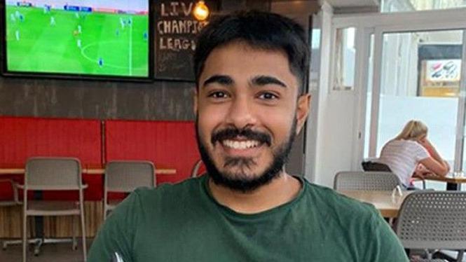 Anugrah Abraham, a man with black hair and facial hair and a dark green T-shirt, smiles directly into the camera. He is sat in a pub, with a TV displaying a football match behind his right shoulder.