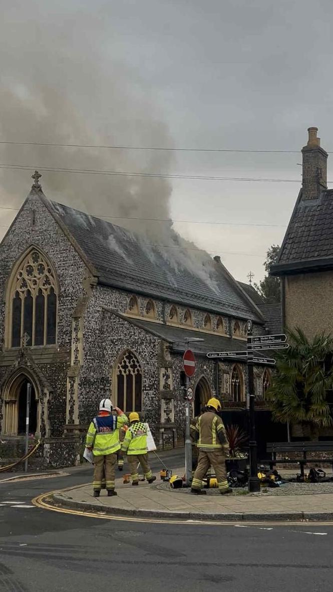 Smoke rising from the tiles on the side of a church roof. There are three firefighters on the pavement on the other side of the road from the church