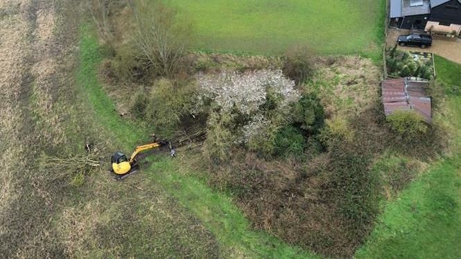 An aerial shot showing an area of woodland and shrubs at the edge of a field which was once a pond