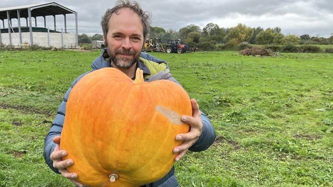 Jonny Crickmore holding a large orange pumpkin up across his torso. He has light brown hair and a short beard, and is wearing a light blue/grey down jacket. Behind him is grass and on the left an open barn structure.
